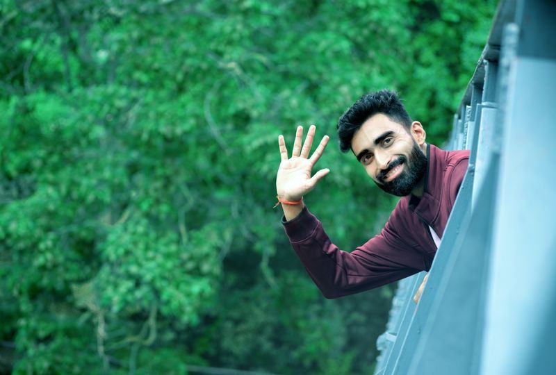 bearded man with smile on face waving hello from bridge with leafy background