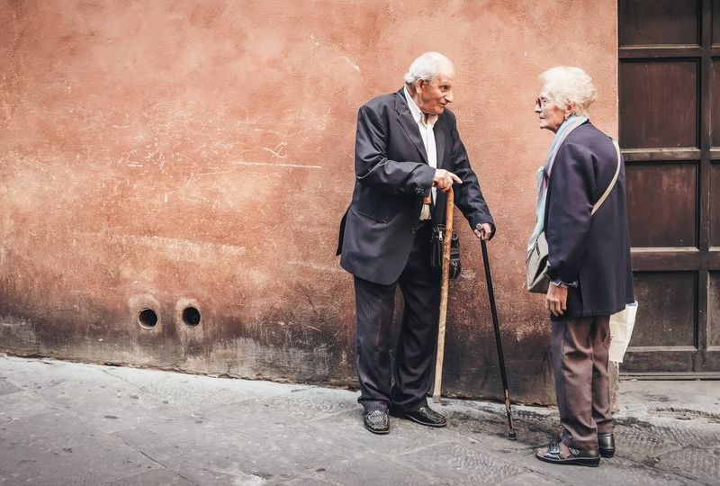 two elderly people talking on street corner with pastel wall and door behind