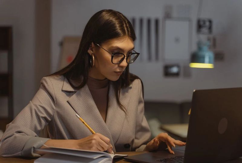 woman with brown hair sitting at a desk in a dark room reading and writing